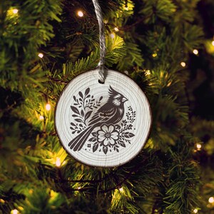 Puede incluir: Un adorno navide&ntilde;o de madera blanco con una silueta negra de un cardenal rodeado de dise&ntilde;os florales.