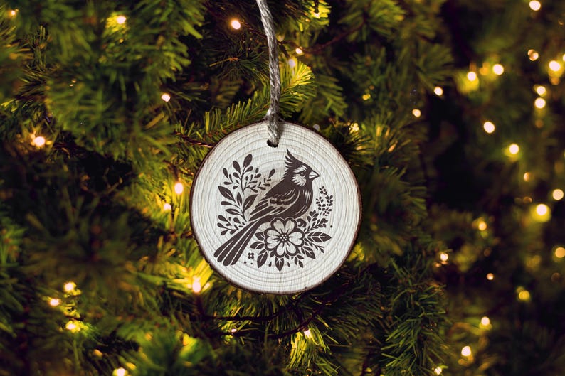 Puede incluir: Un adorno navide&ntilde;o de madera blanco con una silueta negra de un cardenal rodeado de dise&ntilde;os florales.