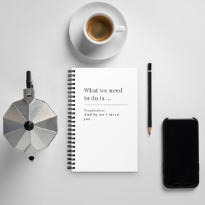 May include: A white spiral bound notebook with the text "What we need to do is... Translation: And by we I mean you."  The notebook is on a white surface with a cup of coffee, a pencil, and a black smartphone.