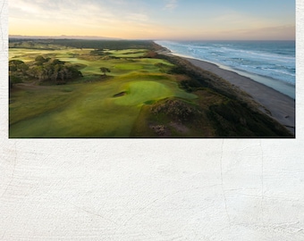 May include: Aerial view of a golf course next to the ocean. The green course contrasts with the sandy beach and blue water. The sky is a gradient of yellow, orange, and blue, suggesting sunrise or sunset. Trees and hills are visible.