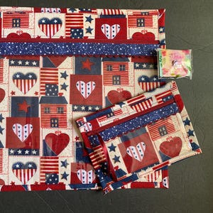 Pode incluir: Um conjunto com tema patri&oacute;tico com uma bolsa grande com z&iacute;per e uma menor. As bolsas s&atilde;o adornadas com um padr&atilde;o repetido de cora&ccedil;&otilde;es, casas, estrelas e bandeiras americanas em vermelho, branco e azul. Um pequeno pingente de bandeira est&aacute; anexado.