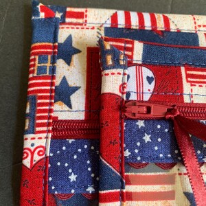 Pode incluir: Close-up de um item de tecido com z&iacute;per vermelho. O tecido apresenta um design patri&oacute;tico com estrelas e listras vermelhas, brancas e azuis. O item &eacute; provavelmente uma pequena bolsa ou estojo, com fecho de z&iacute;per vermelho.