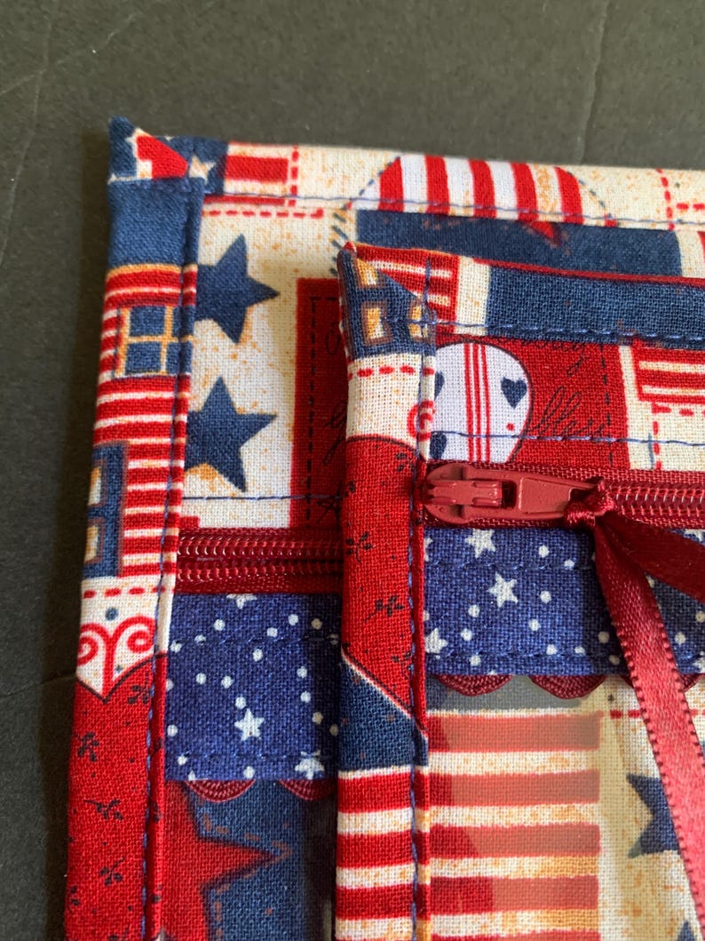 Pode incluir: Close-up de um item de tecido com z&iacute;per vermelho. O tecido apresenta um design patri&oacute;tico com estrelas e listras vermelhas, brancas e azuis. O item &eacute; provavelmente uma pequena bolsa ou estojo, com fecho de z&iacute;per vermelho.