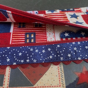 Pode incluir: Uma bolsa com z&iacute;per vermelha, branca e azul com tema patri&oacute;tico. O tecido apresenta padr&otilde;es de estrelas, cora&ccedil;&otilde;es e casas. Uma fita azul com estrelas brancas e uma borda vermelha ondulada adornam a bolsa.