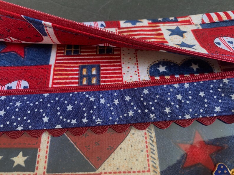 Pode incluir: Uma bolsa com z&iacute;per vermelha, branca e azul com tema patri&oacute;tico. O tecido apresenta padr&otilde;es de estrelas, cora&ccedil;&otilde;es e casas. Uma fita azul com estrelas brancas e uma borda vermelha ondulada adornam a bolsa.