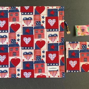 Pode incluir: Um conjunto com tema patri&oacute;tico com um item de tecido retangular grande e uma bolsa menor, ambos adornados com um padr&atilde;o repetido de cora&ccedil;&otilde;es vermelhos, estrelas azuis e casas estilizadas em um fundo creme. O conjunto inclui um pequeno item embalado e um pingente de contas.