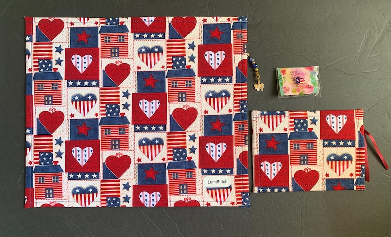 Pode incluir: Um conjunto com tema patri&oacute;tico com um item de tecido retangular grande e uma bolsa menor, ambos adornados com um padr&atilde;o repetido de cora&ccedil;&otilde;es vermelhos, estrelas azuis e casas estilizadas em um fundo creme. O conjunto inclui um pequeno item embalado e um pingente de contas.