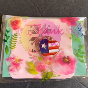 Pode incluir: Um pingente em forma de cora&ccedil;&atilde;o com um design patri&oacute;tico, apresentando listras vermelhas e brancas, um campo azul com uma estrela branca e detalhes dourados. O pingente &eacute; embalado com um cart&atilde;o com tema floral que diz "Thank" e "Lum".