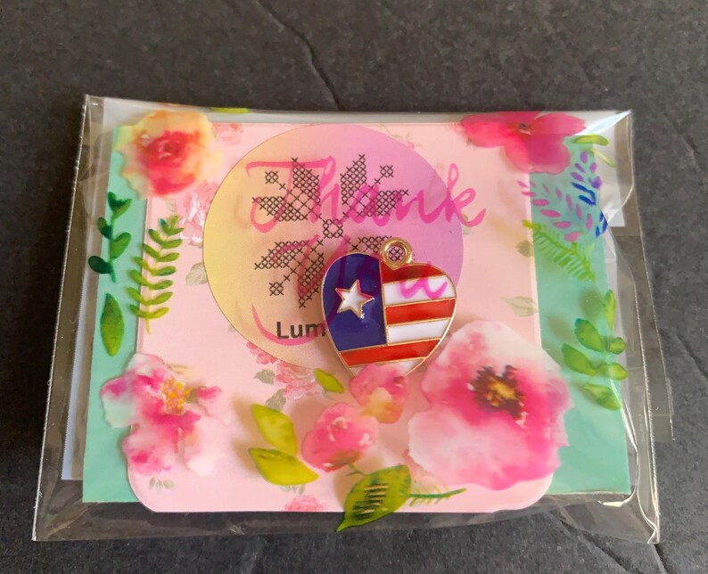 Pode incluir: Um pingente em forma de cora&ccedil;&atilde;o com um design patri&oacute;tico, apresentando listras vermelhas e brancas, um campo azul com uma estrela branca e detalhes dourados. O pingente &eacute; embalado com um cart&atilde;o com tema floral que diz "Thank" e "Lum".