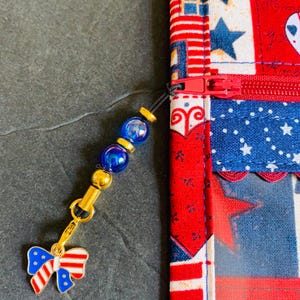 Pode incluir: Uma bolsa com z&iacute;per com tema patri&oacute;tico com z&iacute;per vermelho e um berloque. A bolsa apresenta um design de patchwork com estrelas e listras vermelhas, brancas e azuis. O berloque inclui contas azuis e um la&ccedil;o.