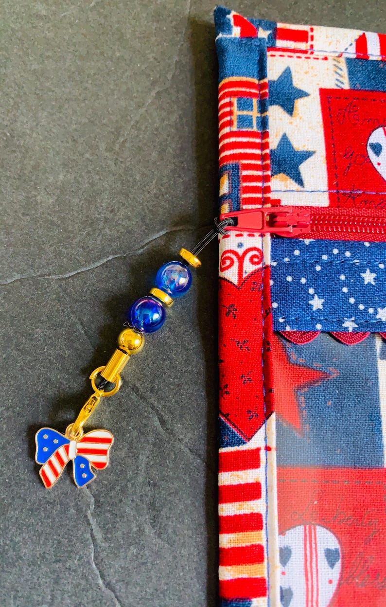 Pode incluir: Uma bolsa com z&iacute;per com tema patri&oacute;tico com z&iacute;per vermelho e um berloque. A bolsa apresenta um design de patchwork com estrelas e listras vermelhas, brancas e azuis. O berloque inclui contas azuis e um la&ccedil;o.