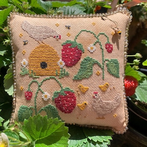 May include: A beige, square, cross-stitched pillow featuring a garden scene. The design includes a yellow beehive, red strawberries, green leaves, a white chicken, and small yellow chicks. The pillow is trimmed with a beaded border.