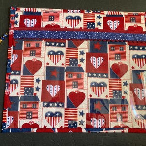 Pode incluir: Uma bolsa com z&iacute;per de tema patri&oacute;tico com um padr&atilde;o repetido de casas, cora&ccedil;&otilde;es e estrelas vermelhas, brancas e azuis. A bolsa tem um bolso de pl&aacute;stico transparente e um z&iacute;per azul com um charme decorativo. O design evoca a bandeira americana.