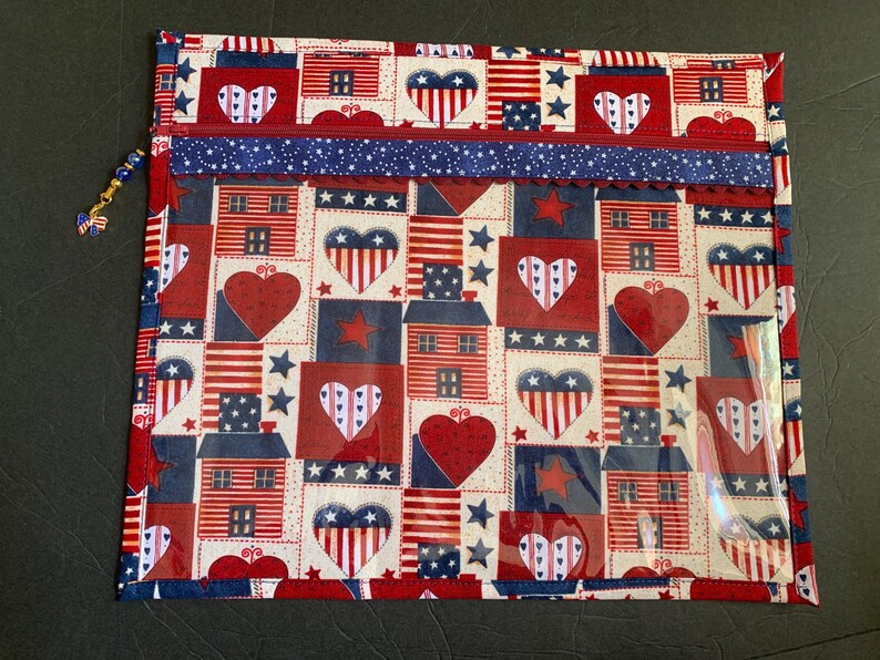 Pode incluir: Uma bolsa com z&iacute;per de tema patri&oacute;tico com um padr&atilde;o repetido de casas, cora&ccedil;&otilde;es e estrelas vermelhas, brancas e azuis. A bolsa tem um bolso de pl&aacute;stico transparente e um z&iacute;per azul com um charme decorativo. O design evoca a bandeira americana.