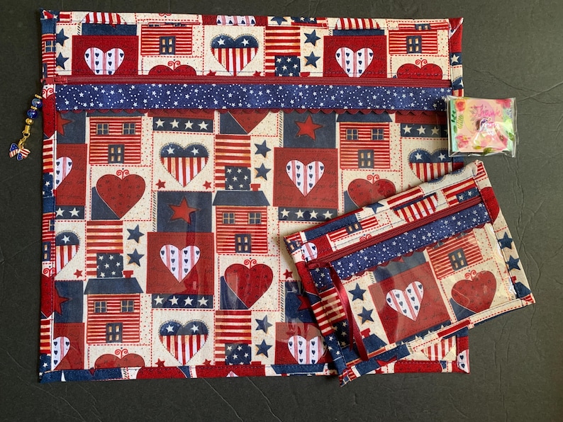 Pode incluir: Um conjunto com tema patri&oacute;tico com uma bolsa grande com z&iacute;per e uma menor. As bolsas s&atilde;o adornadas com um padr&atilde;o repetido de cora&ccedil;&otilde;es, casas, estrelas e bandeiras americanas em vermelho, branco e azul. Um pequeno pingente de bandeira est&aacute; anexado.