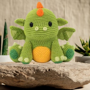 Pode incluir: Um dragão de crochê verde feito à mão com detalhes em laranja. O dragão tem olhos pretos, asas pequenas e uma barriga amarela. Está sentado em uma superfície de pedra. O dragão é um item artesanal caprichoso.