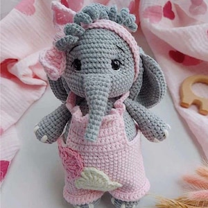 May include: A crocheted gray elephant wearing a pink crocheted dress with a flower design. The elephant has a pink bow on its head.