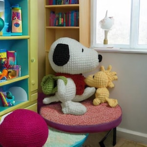 May include: A white crocheted Snoopy doll with a red scarf and a green backpack sits on a purple and pink stool with a yellow crocheted Woodstock doll beside it.