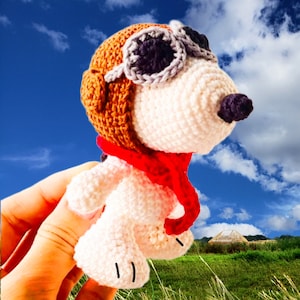 May include: A crocheted stuffed animal wearing a brown aviator hat with a red scarf. The animal has a white body and a brown nose. The animal is holding its head up and looking to the right.