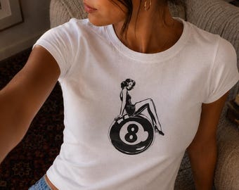 May include: White crop top featuring a black and white graphic of a woman sitting on an eight ball. The shirt has short sleeves and a crew neck. The model is wearing blue jeans.