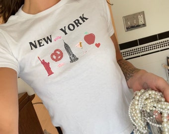 May include: White t-shirt with the text "NEW YORK" and various New York City themed graphics, including the Statue of Liberty, the Empire State Building, a pretzel, a hot dog, a taxi, and an apple. The shirt is being held by a person wearing blue jeans.