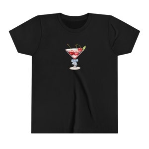 Peut inclure: T-shirt noir avec une illustration &agrave; l'aquarelle d'un verre &agrave; cocktail rempli de liquide rouge et de cerises. Un n&oelig;ud bleu clair est nou&eacute; autour de la tige. Le motif est centr&eacute; sur le devant du t-shirt.