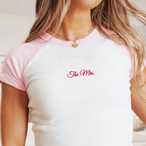 May include: White t-shirt with pink sleeves and neckline. The text "The Mrs." is embroidered in a cursive font on the front. The shirt is a raglan style, with a casual and comfortable design.