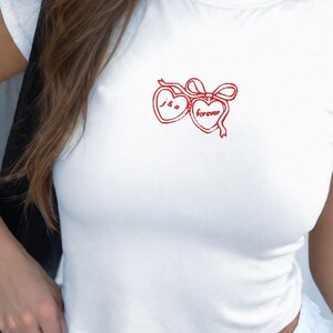 May include: White cropped t-shirt with a red embroidered design of two hearts with a bow. The hearts have the text "J & G" and "forever" inside.