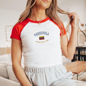 May include: White and red raglan-style t-shirt with the word "VENEZUELA" arched across the chest in blue lettering. Below the word is a small Venezuelan flag and the text "Campe&oacute;n Mundial - 2026".