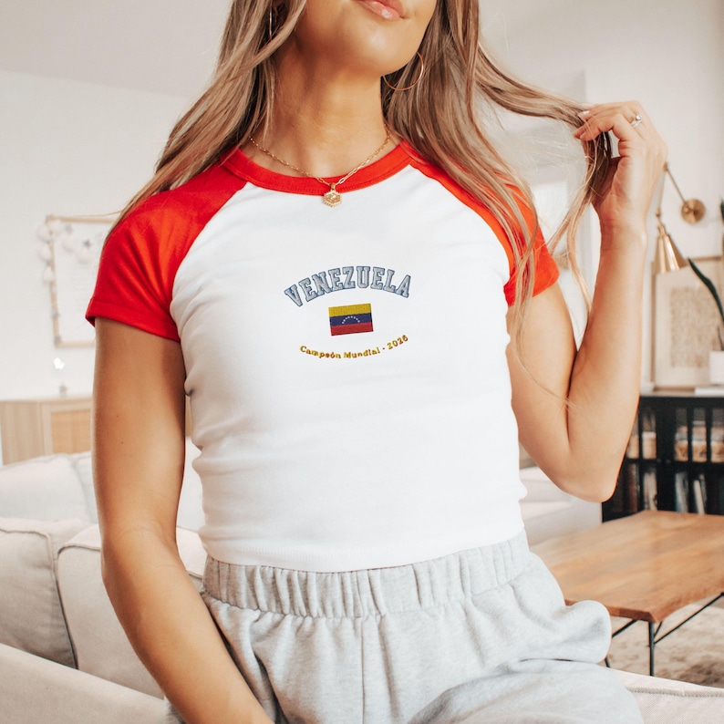 May include: White and red raglan-style t-shirt with the word "VENEZUELA" arched across the chest in blue lettering. Below the word is a small Venezuelan flag and the text "Campe&oacute;n Mundial - 2026".