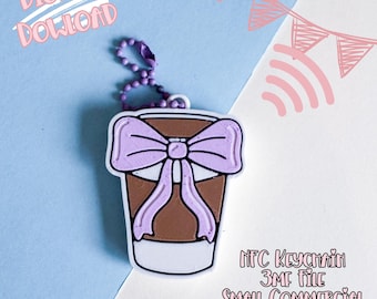 Peut inclure: Un porte-clés en forme de tasse à café avec un nœud violet clair. Le porte-clés est marron et blanc avec une chaîne violette. Le texte sur l'image indique "TÉLÉCHARGEMENT NUMÉRIQUE" et "NFC KEYCHAIN 3MF FILE SMALL COMMERCIAL USE INCLUDED."