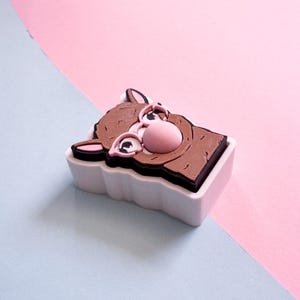 May include: A brown and pink cartoon donkey figure in a white rectangular container. The donkey has large eyes, a pink nose, and pink ears. The background is divided diagonally, with pink and light blue sections.