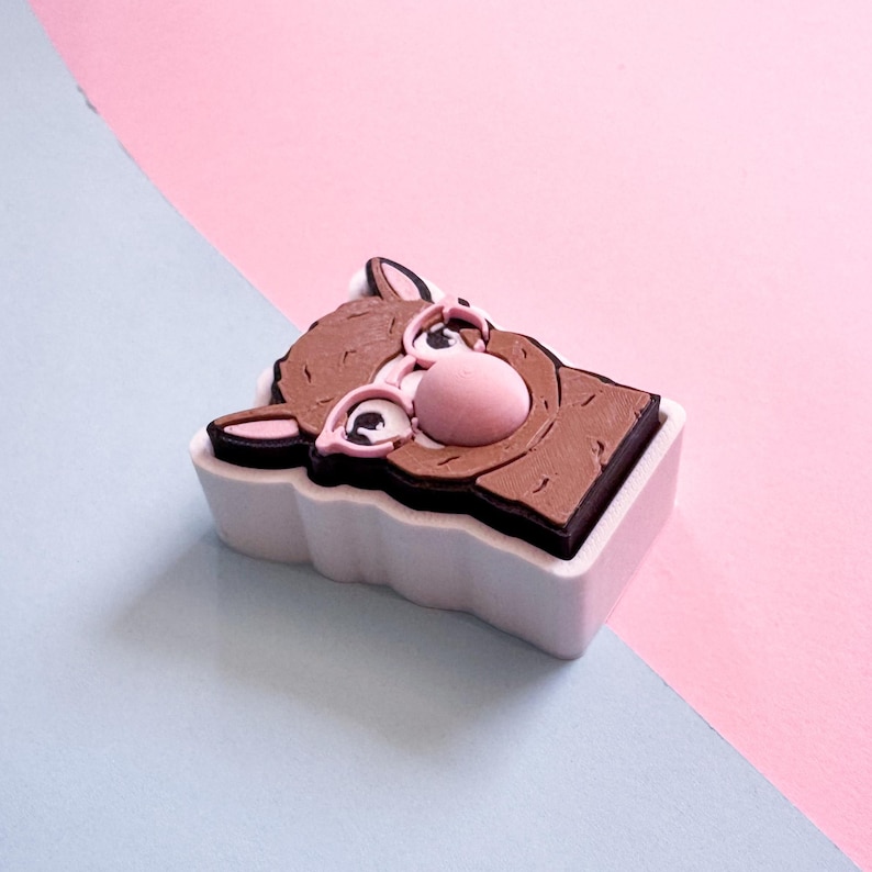 May include: A brown and pink cartoon donkey figure in a white rectangular container. The donkey has large eyes, a pink nose, and pink ears. The background is divided diagonally, with pink and light blue sections.