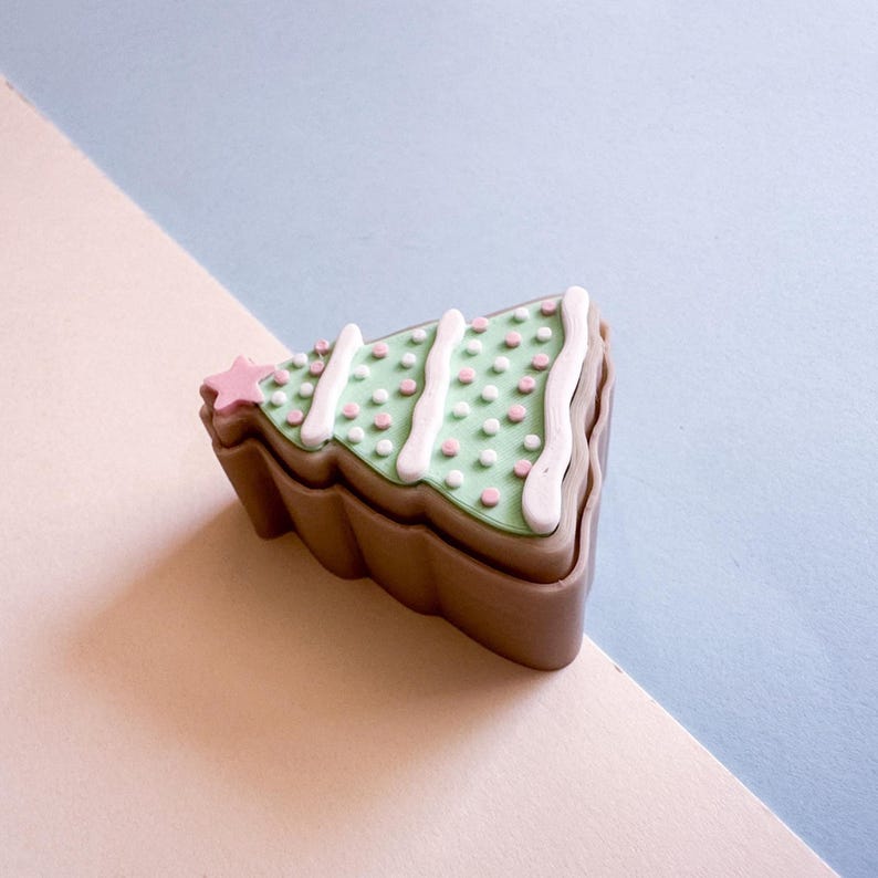 May include: A decorative Christmas tree-shaped box. The box is brown with a green top decorated with white icing stripes and pink and white dots. A pink star adorns the top left corner. The box is set against a light pink and blue background.