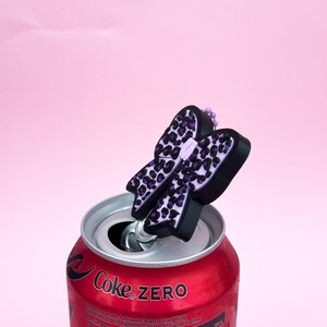 Puede incluir: Una lata roja de Coca-Cola Zero con un adorno para lata en forma de lazo negro y morado. El lazo tiene un dise&ntilde;o de estampado de leopardo. La lata est&aacute; abierta y el adorno est&aacute; sujeto a la pesta&ntilde;a. El fondo es rosa liso.