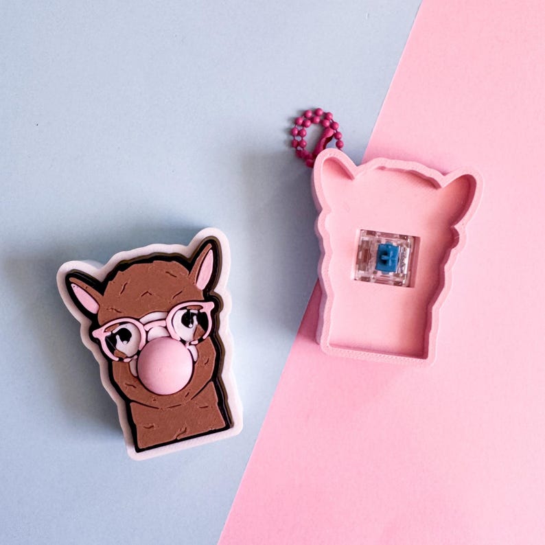 May include: Two llama-shaped keychains. One is brown with pink glasses and a pink nose. The other is a pink, hollow keychain with a small light. The background is split between light blue and pink.