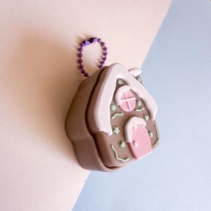 May include: A brown gingerbread house keychain with white frosting accents on the roof and edges. The house features a pink door and window, with green and white decorative details. A purple chain is attached to the top.