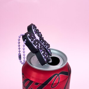 Puede incluir: Un tap&oacute;n de botella con forma de mariposa negra y morada con una cadena de cuentas a juego, apoyado en una lata de refresco roja. El dise&ntilde;o de la mariposa presenta un estampado de leopardo con detalles morados. El fondo es rosa liso.