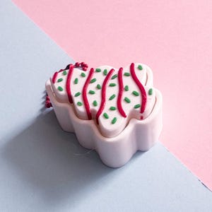 May include: A white, cloud-shaped ornament with red and green decorative accents. The ornament has red lines and green sprinkles on top. The background is split diagonally, with a light blue and pink color scheme.