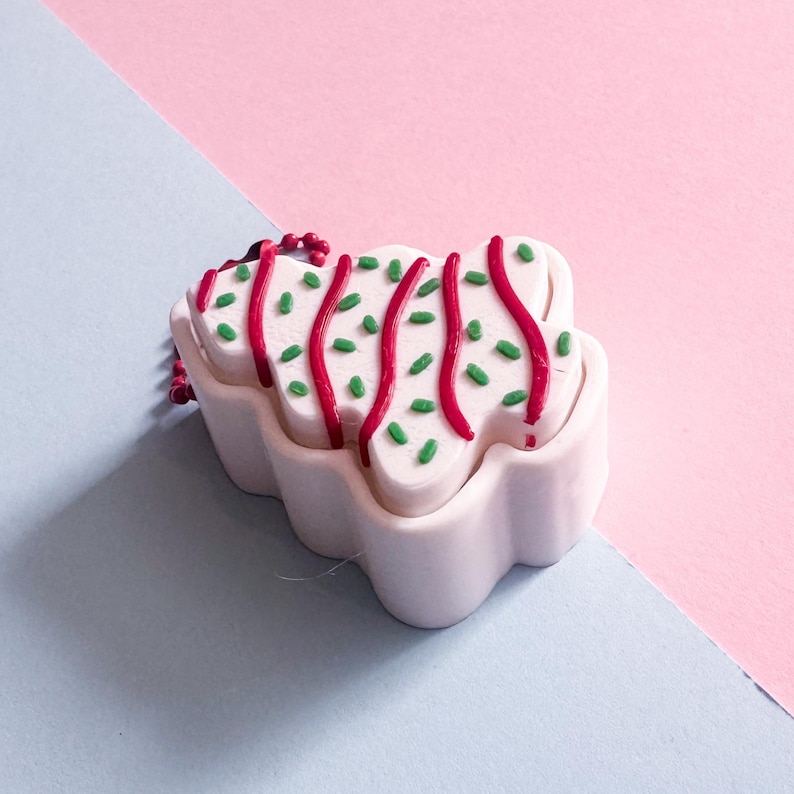 May include: A white, cloud-shaped ornament with red and green decorative accents. The ornament has red lines and green sprinkles on top. The background is split diagonally, with a light blue and pink color scheme.