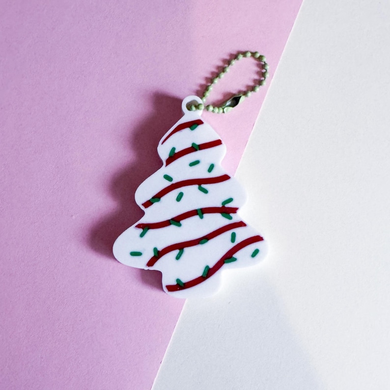 Puede incluir: Un llavero blanco en forma de &aacute;rbol de Navidad con detalles rojos y verdes. El &aacute;rbol est&aacute; decorado con l&iacute;neas rojas y puntos verdes, que simulan luces navide&ntilde;as. El llavero tiene una peque&ntilde;a cadena verde.