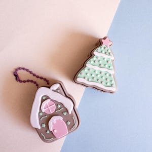 May include: Two Christmas-themed keychains. One is a gingerbread house with pink and white accents, and a purple chain. The other is a green Christmas tree with pink and white polka dots and a pink star on top. Both are on a light pink and blue background.