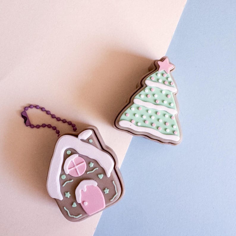 May include: Two Christmas-themed keychains. One is a gingerbread house with pink and white accents, and a purple chain. The other is a green Christmas tree with pink and white polka dots and a pink star on top. Both are on a light pink and blue background.