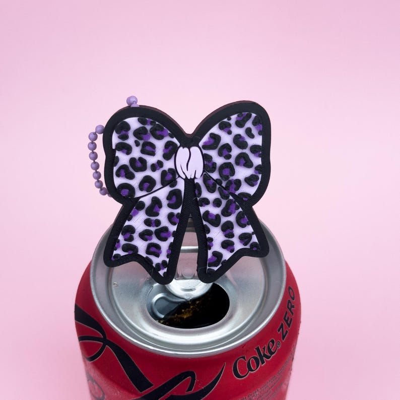 Puede incluir: Un dije en forma de lazo con estampado de leopardo morado y negro, con una peque&ntilde;a cadena, apoyado sobre una lata roja de Coca-Cola Zero. El lazo tiene un contorno negro y un centro morado claro. El fondo es rosa claro liso.