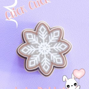 Puede incluir: Un objeto decorativo en forma de copo de nieve con un diseño blanco y gris, sobre un fondo lavanda. La imagen incluye el texto "Click Click" y "Lulu Rabbit", junto con un conejo de dibujos animados sosteniendo un globo en forma de corazón.