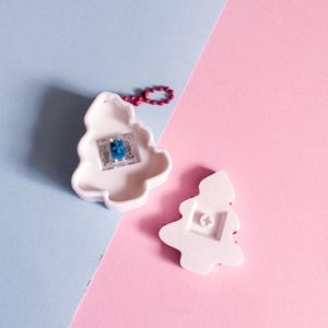 May include: Two white ceramic Christmas tree ornaments. One ornament has a blue and clear component visible, and a red beaded loop. The other ornament is on its side, showing the back. The background is split diagonally, with light blue and pink sections.