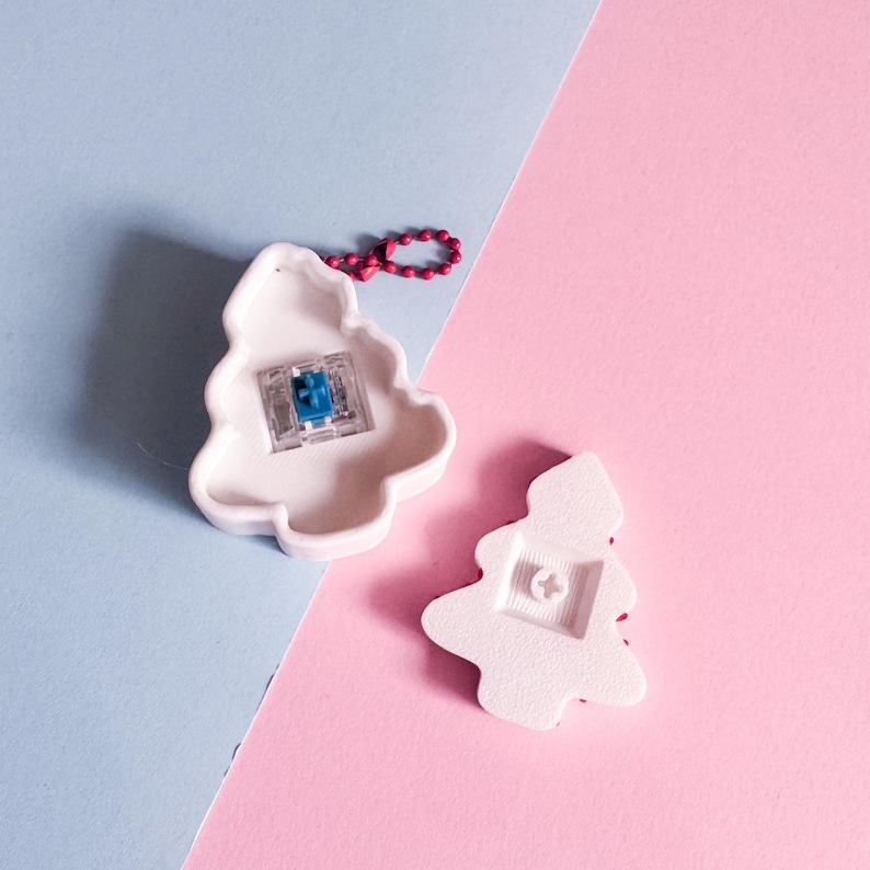May include: Two white ceramic Christmas tree ornaments. One ornament has a blue and clear component visible, and a red beaded loop. The other ornament is on its side, showing the back. The background is split diagonally, with light blue and pink sections.