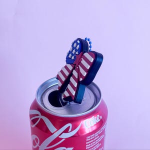 May include: A red Coca-Cola can with a decorative charm featuring a patriotic design. The charm has red and white stripes and blue stars, resembling the American flag. The can is open, with the charm resting on the rim.