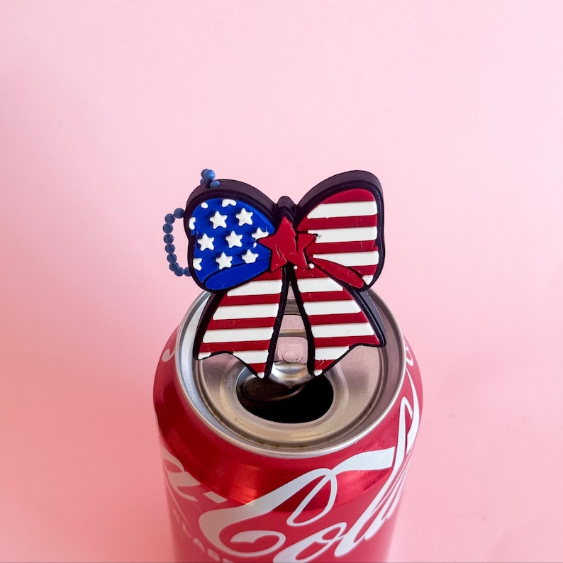 May include: A red Coca-Cola can with a patriotic bow-shaped charm. The charm features the American flag design with red and white stripes, blue stars, and a red center. The background is a solid pink.