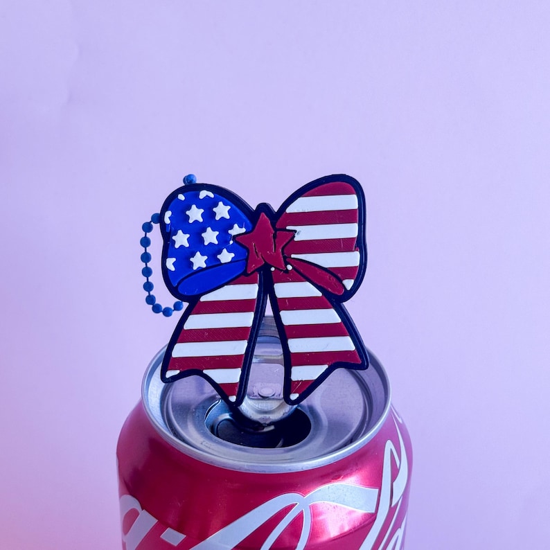 May include: A patriotic bow-shaped charm with a red star and stripes, and blue stars, attached to a chain. The charm is placed on top of a red soda can, against a pink background.